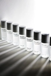 Row of cylindrical translucent bottles with dark caps arranged diagonally across a white surface with strong striped shadows