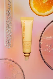Ole Henriksen lip treatment tube in a pastel gradient setting with glass tubes, water droplets, and an orange slice.