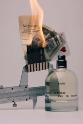 A glass perfume bottle next to a metal caliper holding a burning matchbook, money, and a playing card.