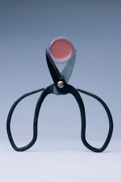 A small circular cosmetic jar held upright in the open blades of black scissors against a light grey gradient background.