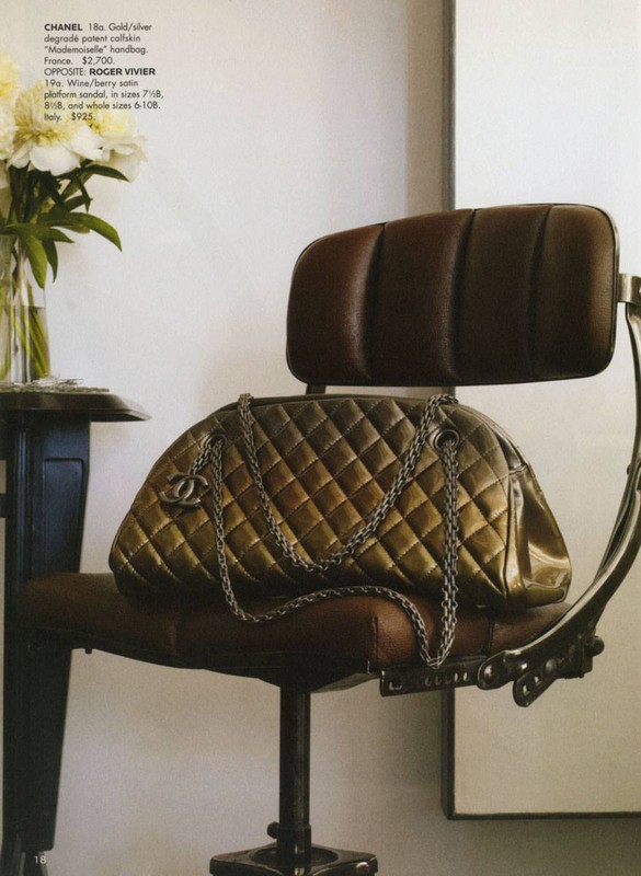 Gold quilted Chanel handbag on a vintage brown rolling chair with silver accents, with a vase of white peonies on a small table.