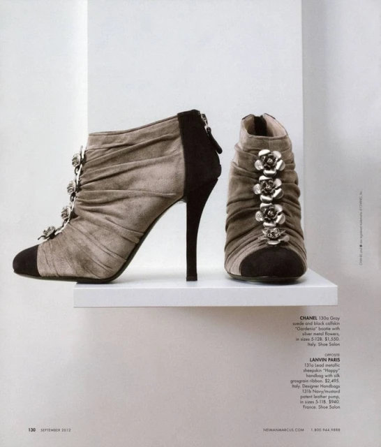 High-heeled ankle boots in gray suede with black accents, decorated with silver flowers, displayed on a white shelf in a magazine shot.