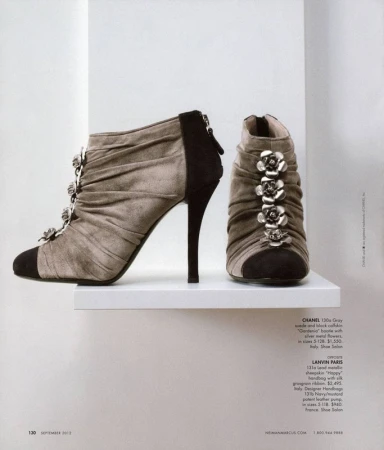 High-heeled ankle boots in gray suede with black accents, decorated with silver flowers, displayed on a white shelf in a magazine shot.