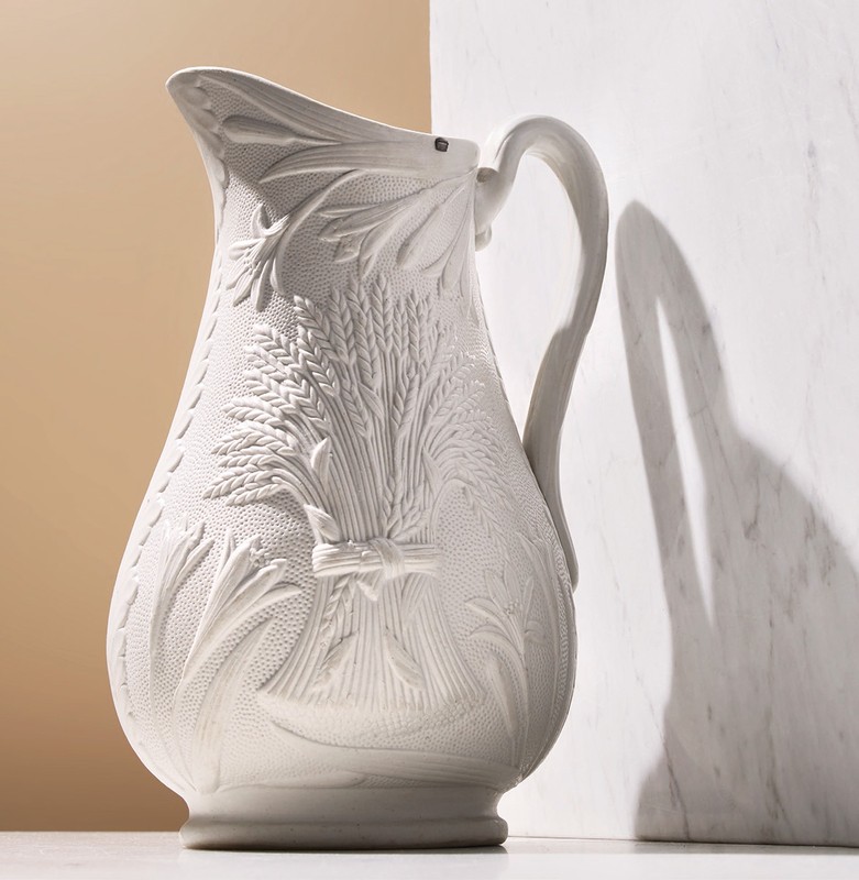 Elegant white ceramic pitcher with wheat motif stands on a marble surface against a two-tone background, casting subtle shadows.