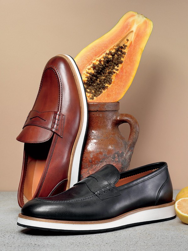 Still life of brown and black leather loafers with platform soles next to papaya and a clay pot against a beige background.