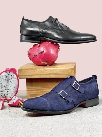 Two dress shoes, a blue suede monk strap and a black cap toe, are displayed with a dragon fruit and a wooden block on a pink backdrop.