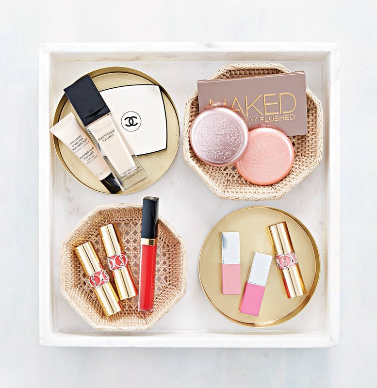 Overhead shot of organized makeup: compacts, foundation, and lipsticks arranged on golden trays within a square white tray.