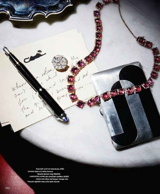 Close-up of jewelry on a white table: necklace, ring, pen, and cigarette case featuring geometric silver and black design.