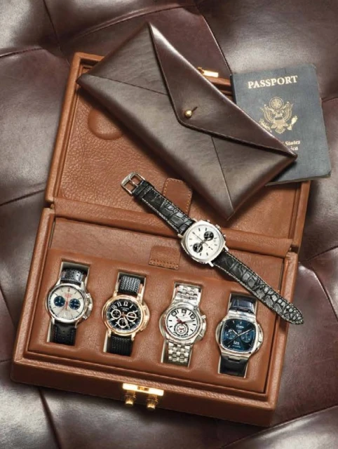 A collection of luxury watches presented in a brown leather box, accompanied by a passport and leather envelope on brown leather.