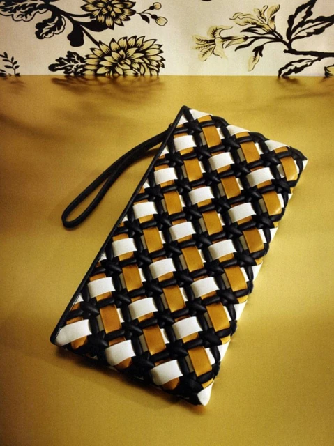 Woven handbag with a black strap on a golden surface with a floral background. The bag is woven with white, black, and tan materials.
