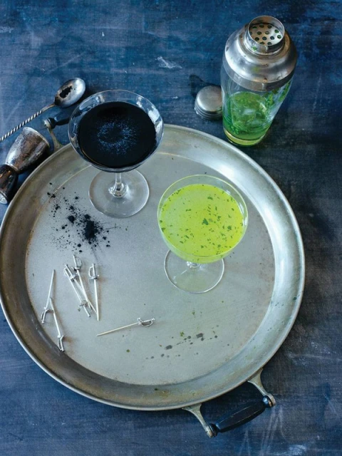 Two cocktails, one black and one green, sit on a silver tray with bar tools and cocktail picks against a dark background, studio lit.