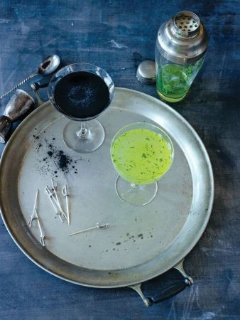 Two cocktails, one black and one green, sit on a silver tray with bar tools and cocktail picks against a dark background, studio lit.