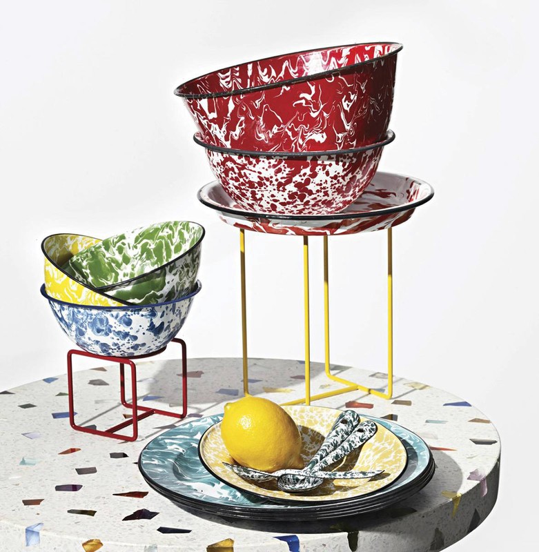 Still life of patterned enamelware bowls and plates stacked on metal stands, arranged around a lemon on a terrazzo surface.