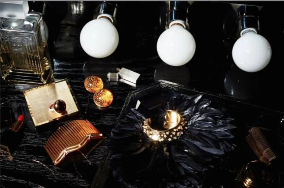 Still life shot of perfume bottle, jewelry, makeup, hair accessories, and decorative items on a glossy black surface with lighting fixtures above.