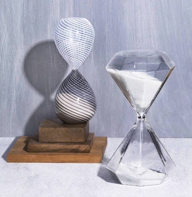 Two hourglasses sit on wooden platforms against a textured gray background. One hourglass is clear with white sand, the other with striped swirls.