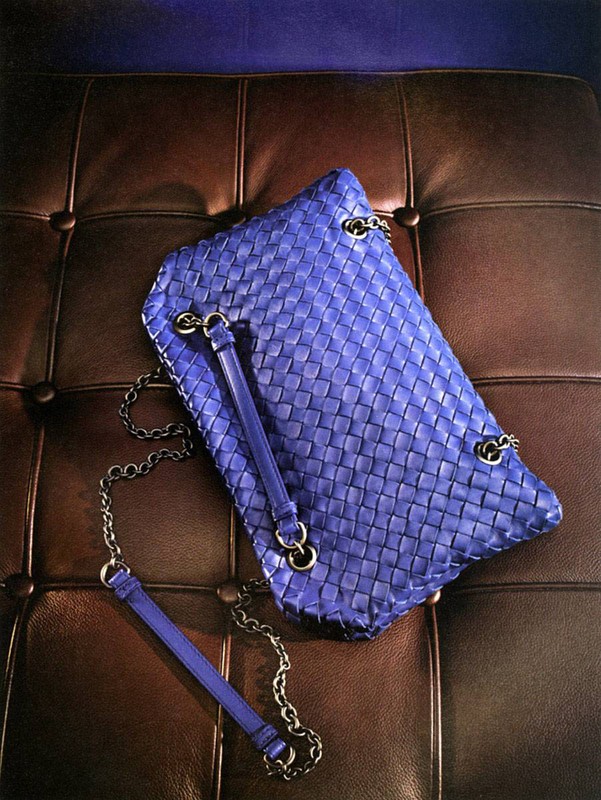 Stylish blue woven shoulder bag with chain strap on a brown leather tufted surface, illuminated by soft light, creating a modern fashion statement.