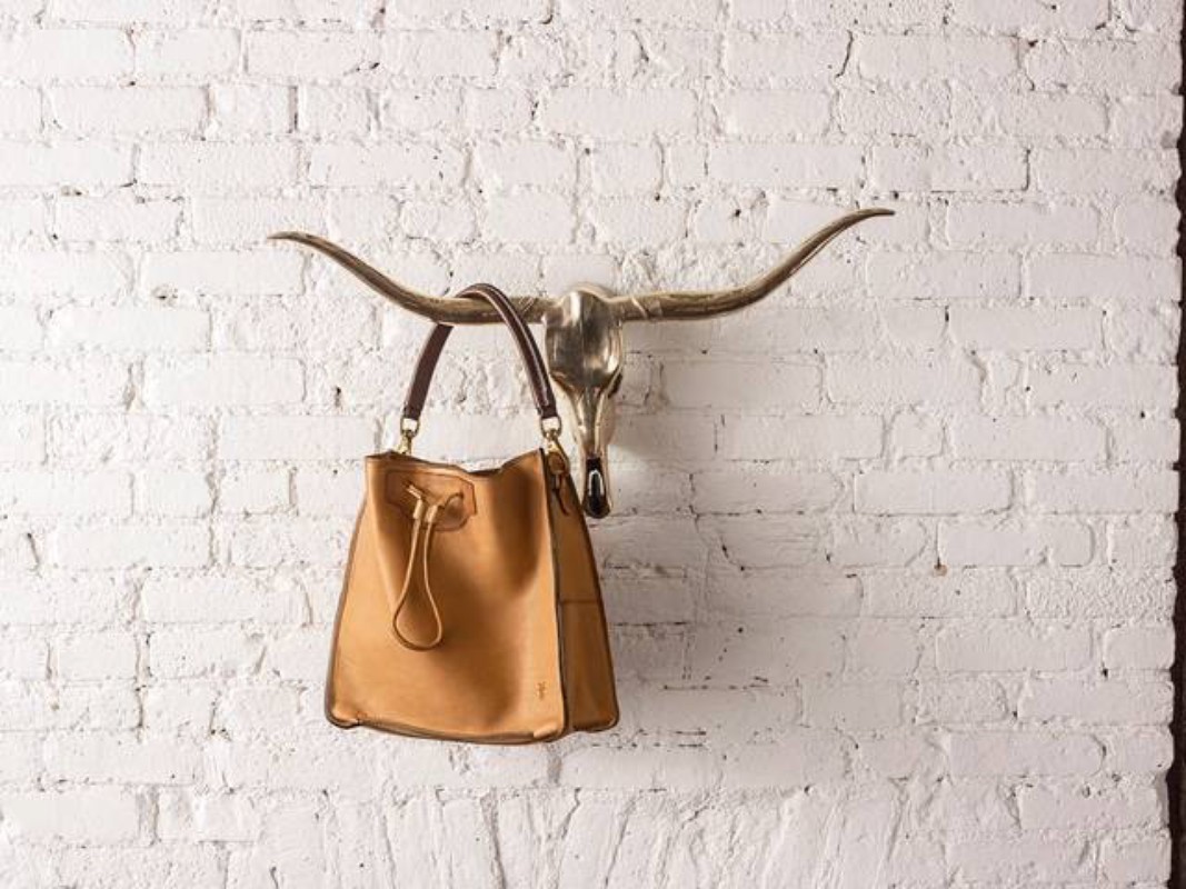 A brown leather bucket bag hangs from a bull skull on a textured white brick wall indoors, a stylish fashion accessory.