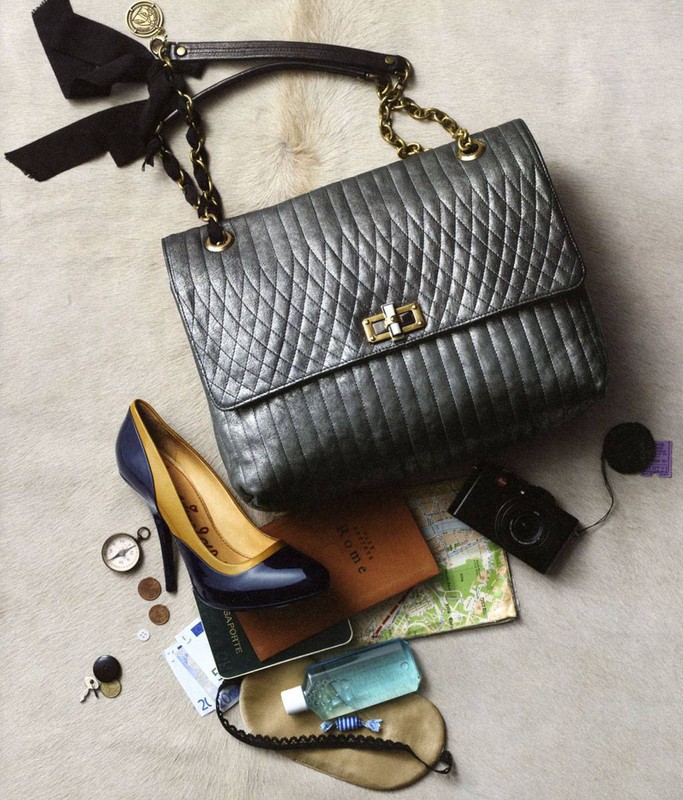 Stylish handbag with a bow, high heel shoe, camera, and travel accessories laid out on a fur-like surface in a still life composition.