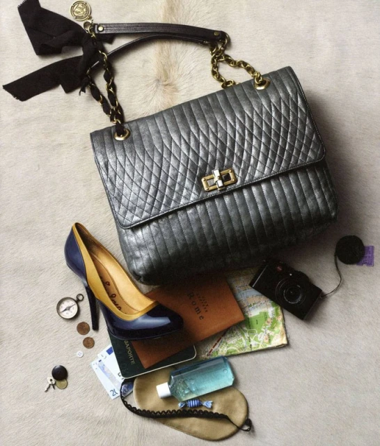 Stylish handbag with a bow, high heel shoe, camera, and travel accessories laid out on a fur-like surface in a still life composition.