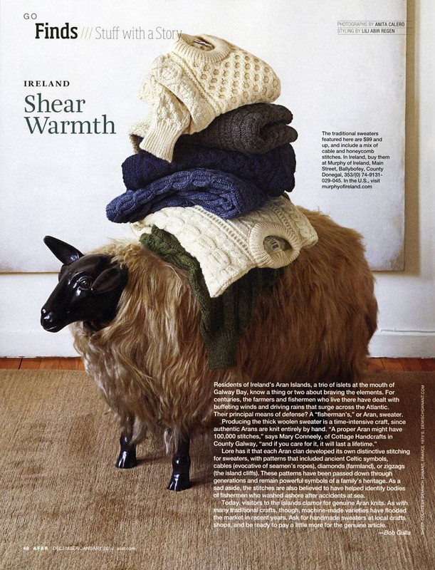 Stacked Aran knit sweaters on a sheep-shaped stool. Knitted Irish wool sweaters in cream, dark grey, and navy, photographed indoors.