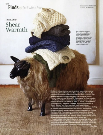 Stacked Aran knit sweaters on a sheep-shaped stool. Knitted Irish wool sweaters in cream, dark grey, and navy, photographed indoors.
