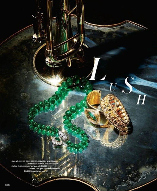 Still life featuring a gold-toned trumpet and emerald and gold jewelry on a reflective surface, showcasing luxury and musical elegance.