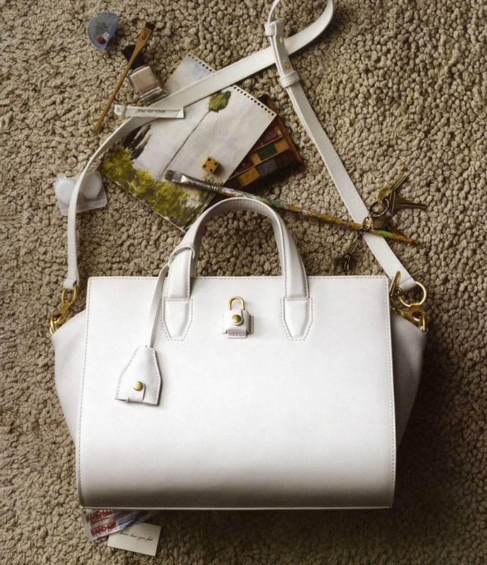 White leather handbag with shoulder strap, artist tools, and paint brushes displayed on textured carpet for a fashion editorial shoot.
