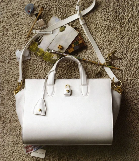 White leather handbag with shoulder strap, artist tools, and paint brushes displayed on textured carpet for a fashion editorial shoot.