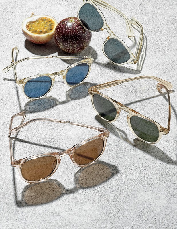 Four pairs of translucent sunglasses are displayed on a gray surface, accented by two passion fruits, one cut open to reveal its pulp.