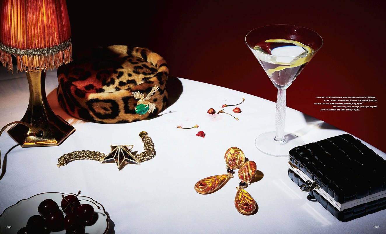 Still life shot of a table with a cocktail, jewelry, cherries, and a leopard print hat against a dark red background.