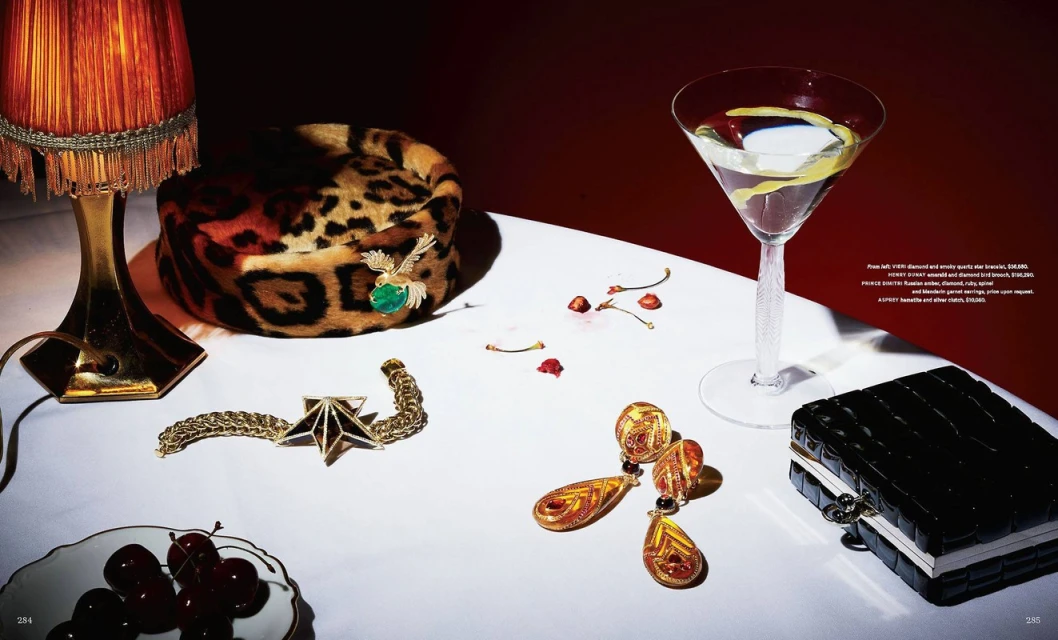 Still life shot of a table with a cocktail, jewelry, cherries, and a leopard print hat against a dark red background.