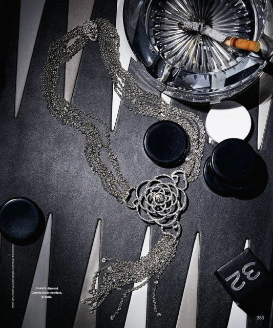 Overhead shot of Chanel diamond Camelia Brode necklace, backgammon pieces, cigarette in crystal ashtray, and black and silver board.