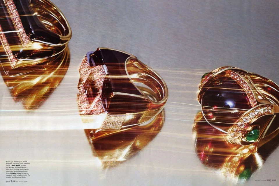 Three gold statement rings with gemstones and diamonds are showcased on a reflective surface with light streaks and shadows.