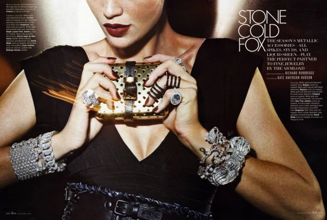 Close-up fashion shot: woman poses with heavy jewellery, gold clutch, and dark lipstick against soft-lit backdrop. Metallic accessories vogue.