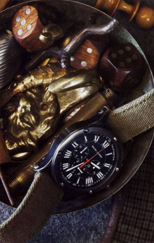 Overhead shot of a black wristwatch with a beige canvas strap nestled in a metal bowl amongst dice, a gold lion head, and other trinkets.