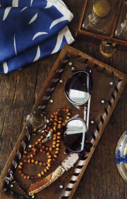 Still life of sunglasses, amber prayer beads, and pocket knife on a decorative tray resting on rustic wooden planks, next to an antique box.
