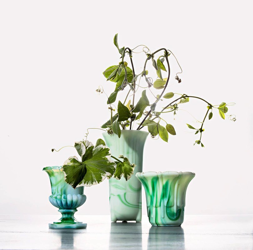 Still life composition featuring three variegated green glass vases of different shapes holding leafy green sprigs against a white background.