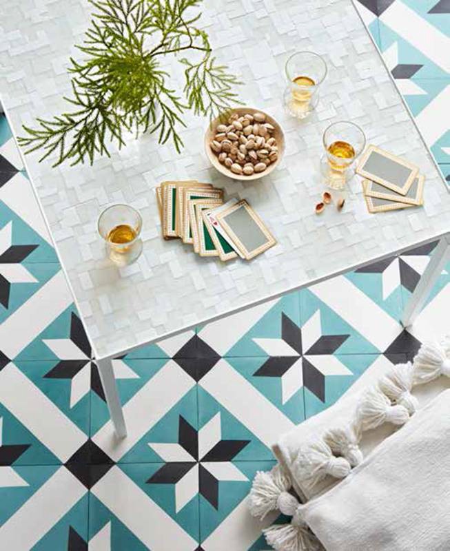 Overhead shot of a square table with drinks, snacks, plants, and cards, set on a geometric-patterned floor with a white throw pillow.