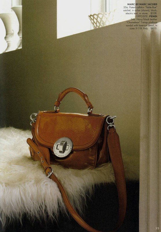 Tan leather satchel handbag with silver hardware and shoulder strap, styled on a white faux fur surface against a neutral wall in a fashion editorial.