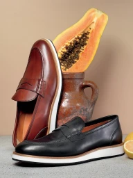 Brown leather loafer and black leather loafer posed with a halved papaya, ceramic jug, and lemons on a neutral surface.
