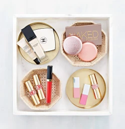 Flat lay of luxury beauty products arranged on gold trays and woven baskets inside a white marble tray.