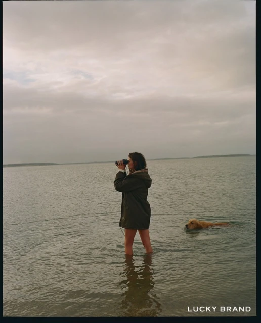 A person stands knee-deep in water at a beach looking through binoculars with a dog swimming nearby, taken on an overcast day.