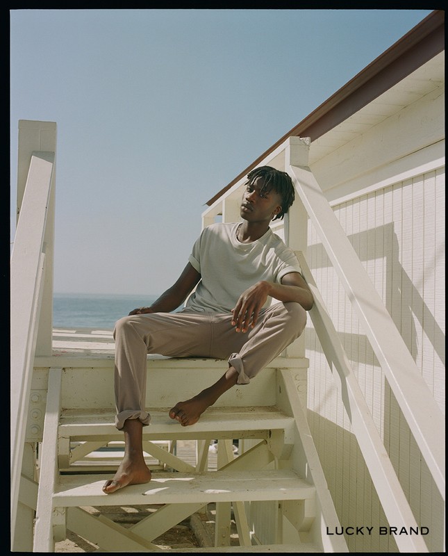 Young man sits on white wooden beach stairs wearing a t-shirt and khakis with ocean and blue sky in background on sunny day.