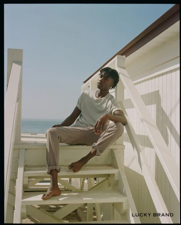 Young man sits on white wooden beach stairs wearing a t-shirt and khakis with ocean and blue sky in background on sunny day.