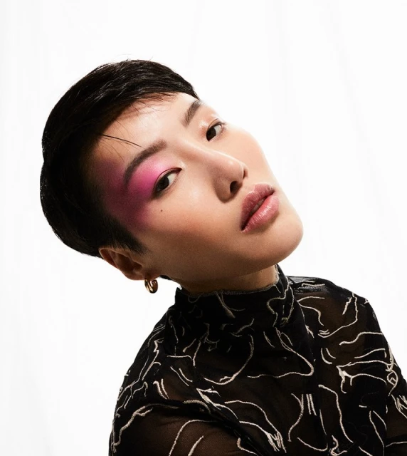 Close-up portrait of a young Asian woman with a pixie cut, wearing a black patterned top, against a bright white background.