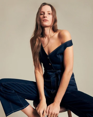 Model seated on a stool wearing a dark denim jumpsuit with contrast stitching, styled by Lindsay Grosswendt.