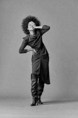 Smiling woman with afro hairstyle wearing a ruched midi dress and tall leather boots covering her eyes with one hand