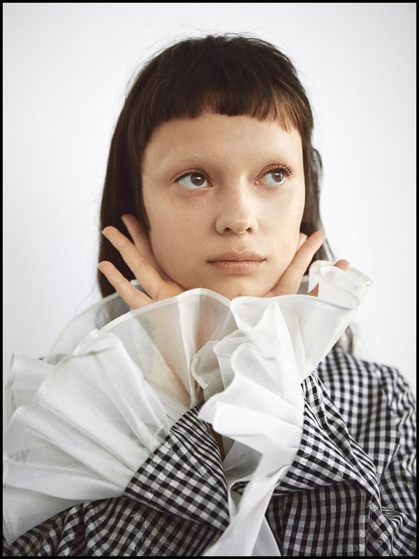 Close-up portrait of a young adult female with dark hair and pale skin, wearing a black and white checkered top with ruffled collar against a white background.