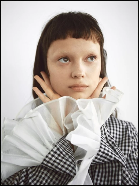 Close-up portrait of a young adult female with dark hair and pale skin, wearing a black and white checkered top with ruffled collar against a white background.