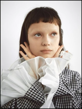 Close-up portrait of a young adult female with dark hair and pale skin, wearing a black and white checkered top with ruffled collar against a white background.